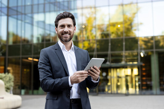Professional businessman wearing a suit and white shirt smiling at the camera while holding a digital tablet with both hands, standing outdoors in front of a modern glass building