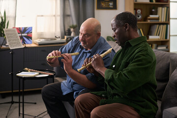 Middle aged Caucasian man and middle aged Black man sitting on sofa practicing playing wooden flutes together, focusing on finger placement, music stand with sheet music in background © pressmaster