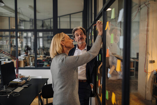 Colleagues looking at sticky notes at glass pane in office