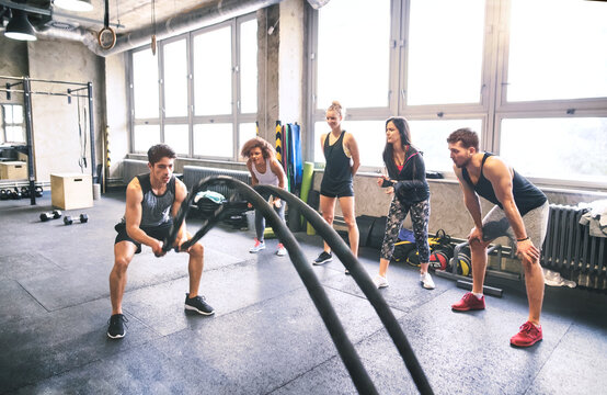Group of young fit people cheering at man exercising with ropes in gym