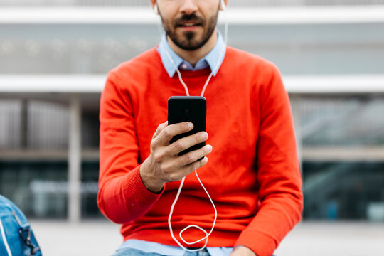 Casual businessman sitting on a bench in the city, using his smartohone and earphones