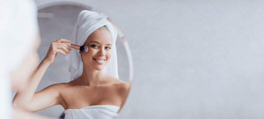 A woman stands in front of a mirror putting on makeup. She is smiling as she uses a brush on her face, wrapped in a towel. The setting shows a simple bathroom with soft light. © Prostock-studio