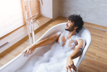 A man sits in a bathtub filled with bubbles. Sunlight comes through the window, and the bathroom has a modern design with wooden floors. He appears to enjoy a moment of relaxation.