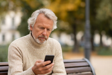 Obraz premium senior man in casual sweater sitting on park bench, anxious expression while looking at phone, autumn city park background softly blurred, natural outdoor lighting, candid composition