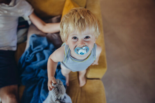Little boy with pacifier standing on couch, looking up at camera