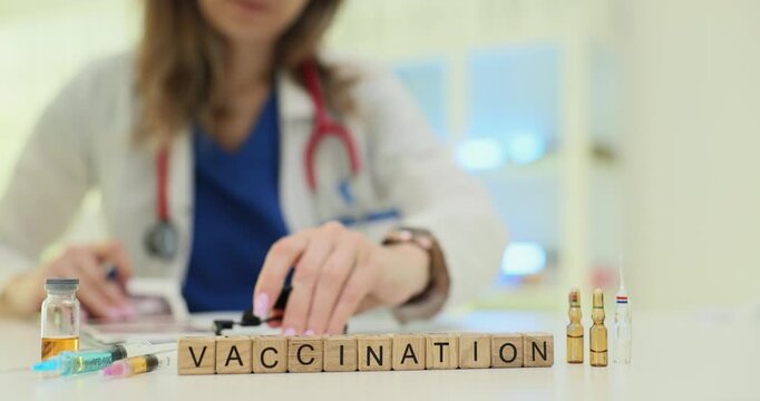 Wooden blocks spell word Vaccination near syringes and vials on clinic desk. Lady doctor arranges ampoules preparing shot supplies for immunization