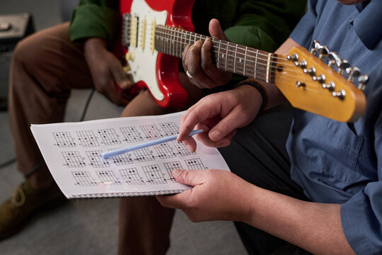 Middle aged Black man playing electric guitar while middle aged Caucasian man holding chord chart and pointing at diagrams, both sitting closely during music lesson or practice session