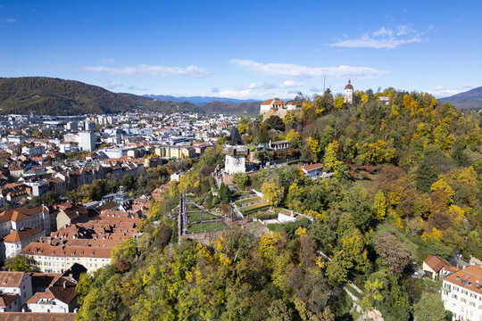 Aerial view of the Schlossberg hill crowned with the iconic clock tower, overlooking the old town's terracotta rooftops and the distant, hazy mountains, Graz, Styria, Austria.