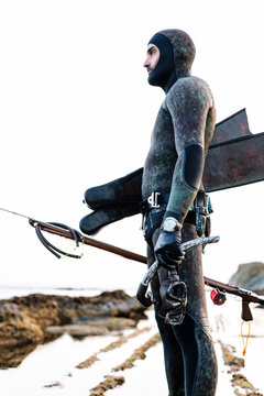 Man wearing wetsuit holding scuba mask and harpoon while standing at beach against clear sky