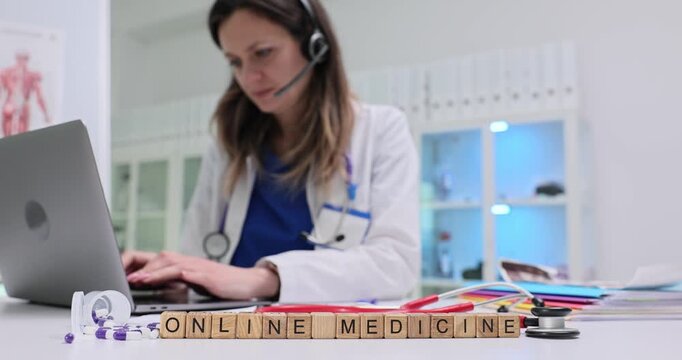 Wooden blocks spell phrase Online Medicine near laptop on clinic desk. Lady doctor types on keyboard answering remote consult while headset stays on