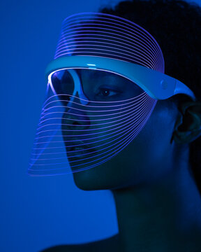 Close-up of young woman wearing blue led mask against wall at home