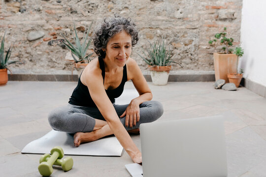 Smiling woman using laptop for learning yoga through online tutorial while sitting at back yard