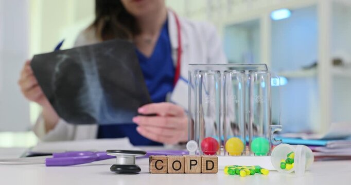 Wooden blocks spell abbreviation COPD on clinic desk. Doctor holds chest X-ray while reviewing respiratory results near spirometry tubes in clinic