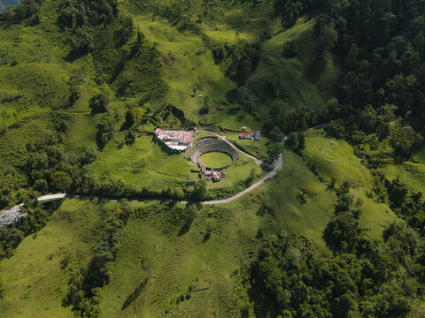Aerial view of a circular arena nestled amidst rolling green hills and verdant foliage, a stark contrast to the terracotta rooftops, Salento, Quindio, Colombia.