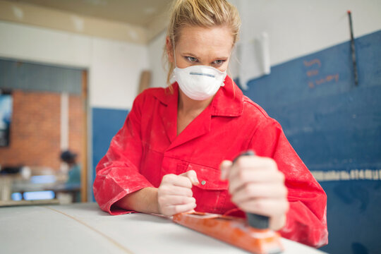 Surfboard shaper workshop, female surfshop employee wearing overalls and mask sanding surfboard
