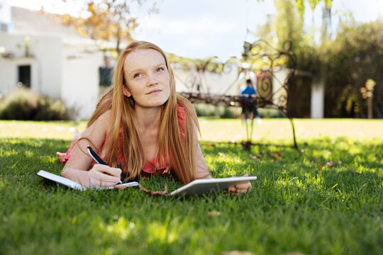 Grl with long red hair lying in grass with tablet and notebook