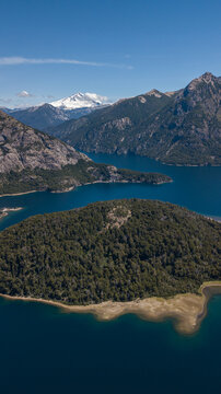Aerial view of an island covered in lush green trees surrounded by deep blue waters, with snow-capped mountains in the background, San Carlos de Bariloche, Rio Negro Province, Argentina.