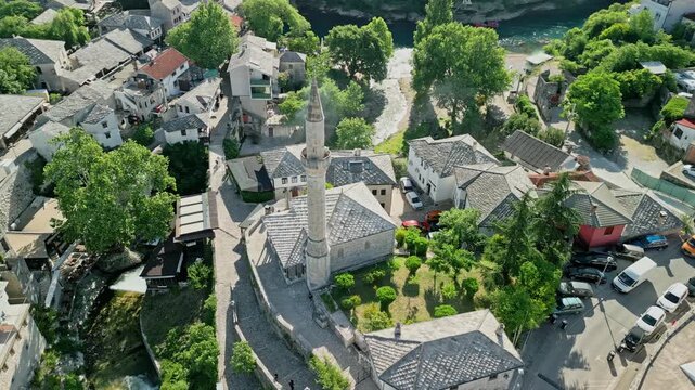 Mostar, Bosnia and Herzegovina - 05 28 2025: Arc drone video of Nezir Agina Dzamija with Crooked Bridge and Radobolja River, Neretva River and Hajji Kurts Mosque in Mostar Old Town on a sunny day.