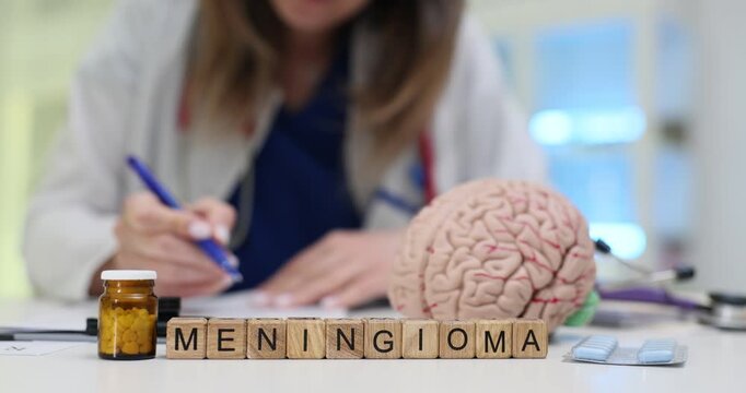 Wooden blocks spell word Meningioma near brain model and pills on clinic desk. Doctor points at cortex model planning imaging tests for tumor case