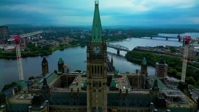 The cloudy sky over Ottawa's Parliament Hill clock tower with the Canadian flag gently waving captured from an aerial drone during renovation work and construction vehicles parked around building T1-2