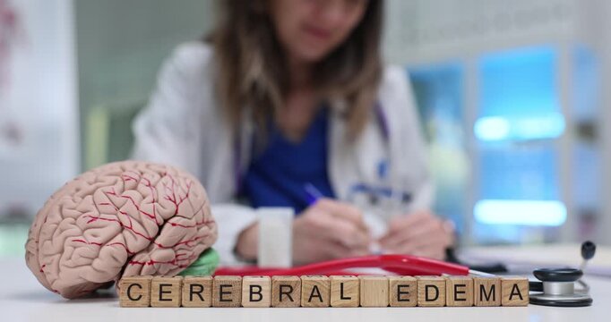 Wooden blocks spell phrase Cerebral Edema near brain model on clinic desk. Doctor writes notes checking neurologic swelling signs of patient carefully