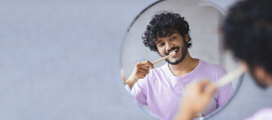 A person stands smiling while brushing teeth in front of a round mirror. The light fills the bathroom. The individual holds a toothbrush and shows good hygiene. © Prostock-studio