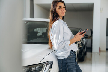 Smartphone is in hands, leaning on the vehicle. Woman posing with a car in an auto showroom, modern dealership environment