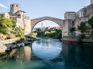 Obraz premium Mostar, Bosnia and Herzegovina 05 28 2025: Frontal drone photo of Old Bridge Mostar in Old Town with Halebija Tower, Sultan Selimov Mesdžid, Koski Mehmed Pasha Mosque, Neretva River, boats, and Beach