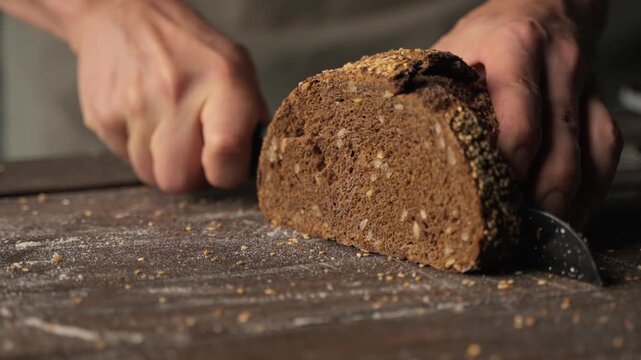 Baker slicing a fresh loaf of rustic whole grain rye bread on a wooden cutting board with a bread knife