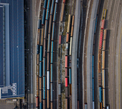 Aerial view of a symphony of steel and shadows as trains rest on converging tracks, a modern ballet of industry unfolds beside solar panels, Chiasso, Ticino, Switzerland.