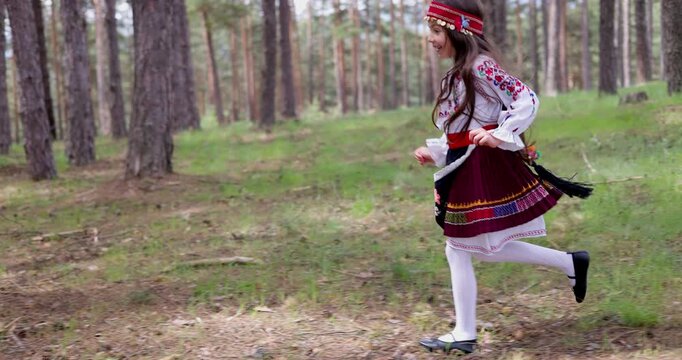 Little girl with ethnic bulgarian folklore costume running behind trees on a trail in forest, Bulgarian beautiful nature