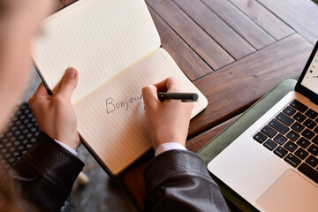 High angle view of a person writing the word "Bonjour" in a notebook with a pen. A laptop and a green sleeve sit on a wooden table. © dsbedarieva
