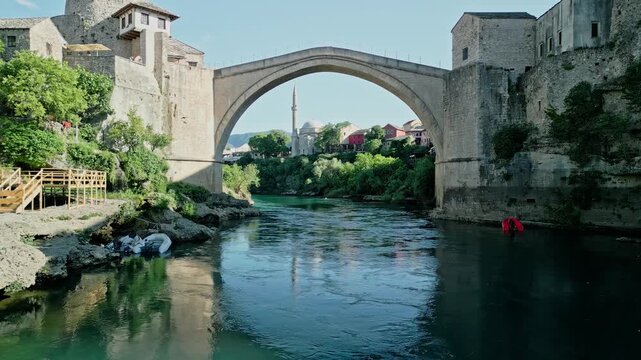 Mostar, Bosnia and Herzegovina - 05 28 2025: Frontal drone video of Old Bridge Mostar in Old Town with Halebija Tower, Sultan Selimov Mesdžid, Koski Mehmed Pasha Mosque, Neretva River, boats, and bird