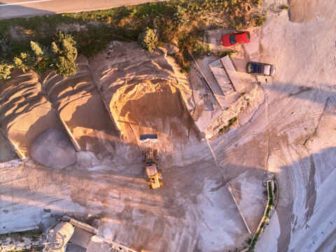Aerial view of a quarry revealing contrasting textures of sand and stone, a lone wheel loader, and parked cars near the edge, Manilva, Andalusia, Spain.
