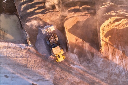 Aerial view of a colossal wheel loader amidst the stark, textured landscape of a quarry, plumes of dust rising in the golden light, Manilva, Andalusia, Spain.