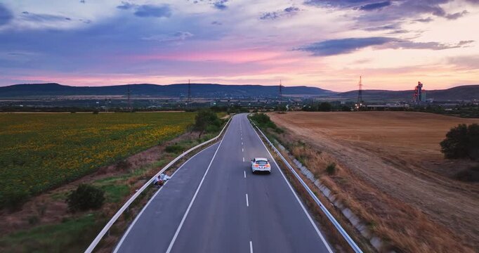 Aerial View of Fast Sports Car Driving Along Country Road Between Sunflower Field and Wheat at Sunset