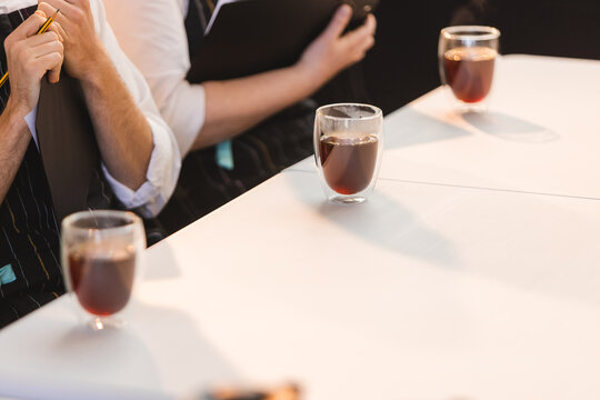 Professional coffee cupping session with judges taking notes. Three double-walled glass cups with black specialty coffee on a white table. Baristas evaluating brew quality in a bright, modern cafe.
