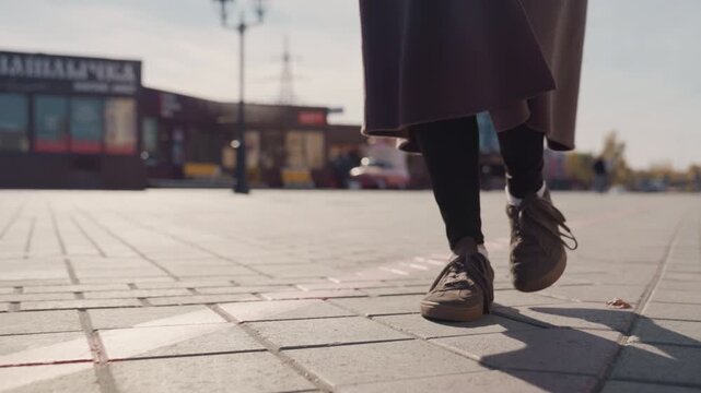 Low angle walking shoes on pavement, coat hem swaying, sunlight casting long shadows across tiled plaza, slow purposeful steps, urban storefronts blurred in background, casual sneakers scuffing