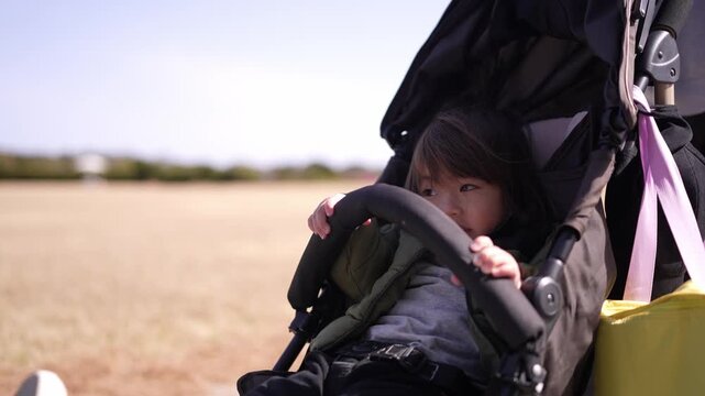 A young girl sits in a stroller with a pink bag hanging from the handle. The scene is peaceful and calm, with the girl looking out at the grassy field
