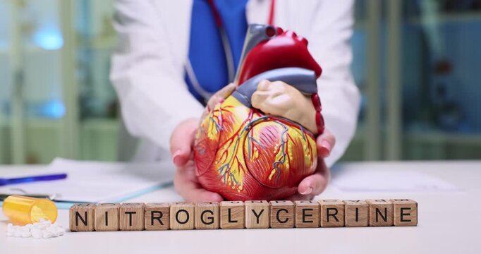 Miniature wooden cubes on desk form word Nitroglycerine by tablets. Woman cardiologist holds anatomical model of human heart in hands showing students