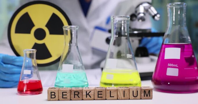 Lab worker shows radiation warning sign to emphasize potential danger. Wooden cubes on table form word Berkelium indicating substance under analysis