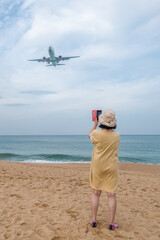  Solo travel in old age gray travel or sinior tourism - an elderly active woman photographs a low-flying plane in the early morning at Mai Khao Planes beach Phuket in Thailand