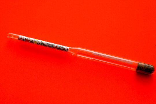 A glass hydrometer for measuring alcohol strength is placed horizontally on a rich red background. The device features a clear color-coded scale for precise determination of alcohol concentration in h