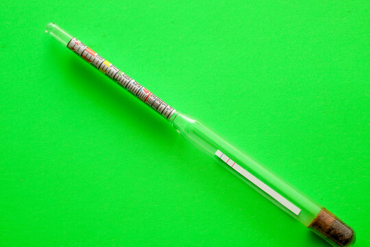 A glass household alcoholometer is placed diagonally on a vivid light green background. This measuring device is used to determine the strength of drinks and control quality in home production.