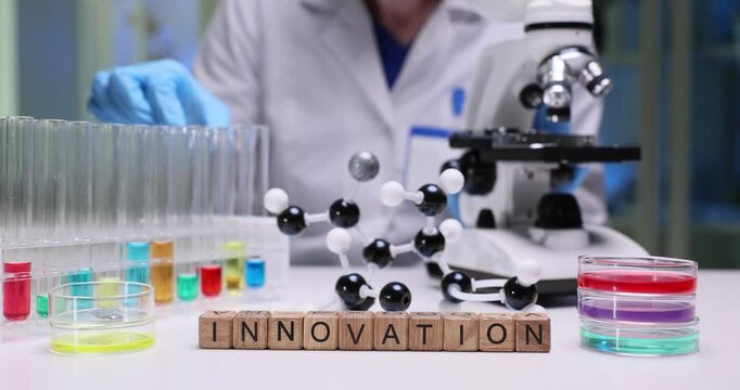 Laboratory worker carefully examines different liquids inside clear test tubes. Miniature wooden cubes on table form word Innovation by molecule model