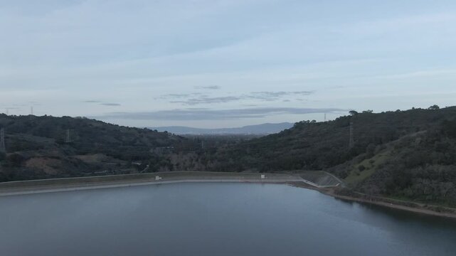 A cinematic wide aerial shot overlooking the calm waters of Steven Creek Reservoir nestled in the Santa Cruz Mountains. The footage captures the surrounding green hills and the concrete dam structure