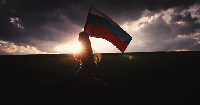 Bulgarian girl waving bulgarian flag and run against dramatic sunset sky on a green field, concept for freedom spirit, bulgarian independence and liberation