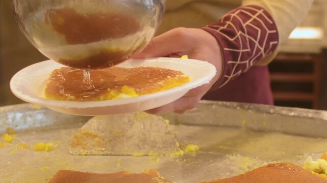 Close up kunafa dessert being served in paper plate with hot sugar syrup poured from metal bowl street sweets shop food preparation close up. Popular middle east and mediterranean dish
