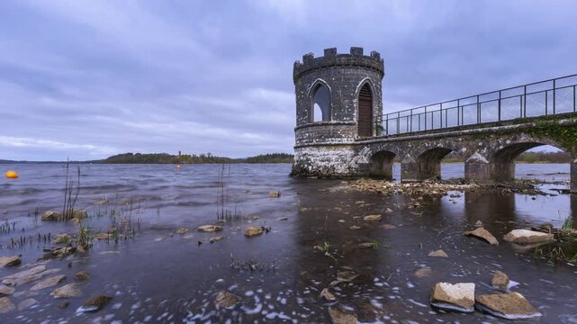 Timelapse of a medieval tower on lake with shoreline rocks in the foreground and medieval castle in the distance on a cloudy day in county Roscommon in Ireland.