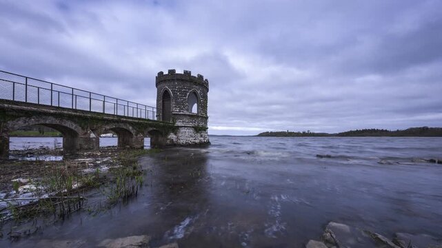 Timelapse of a medieval tower on lake with shoreline rocks in the foreground and medieval castle in the distance on a cloudy day in county Roscommon in Ireland.